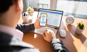 Mężczyzna zatwierdza przelew bankowy na telefonie, korzystając z bankowości online na laptopie.