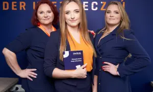 Trzy kobiety w granatowych marynarkach i pomarańczowych bluzkach pozują do zdjęcia. Jedna z nich trzyma broszurę Unilink.