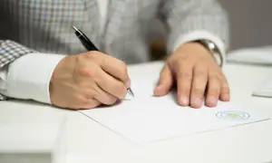 Mężczyzna w eleganckim garniturze pisze długopisem na białej kartce. Widoczny jest wzór czynnego żalu na dokumencie.