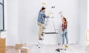 Para odnawia mieszkanie. Mężczyzna na drabinie maluje ścianę, kobieta obok też maluje.