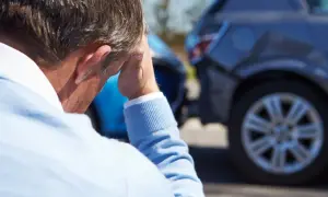 Mężczyzna w niebieskim swetrze z ręką na głowie, na tle uszkodzonych samochodów.