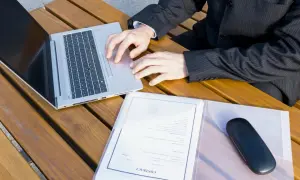 Biznesmen pracuje na laptopie przy drewnianym stole, obok dokumenty z nagłówkiem "CONTRACT". Przygotowuje wpis do księgi wieczystej.