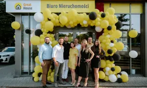 Grupa ludzi pozuje przed biurem nieruchomości ozdobionym żółtymi balonami.
