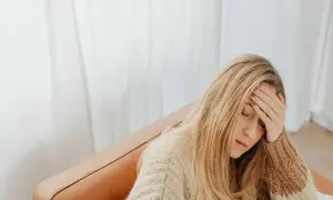 Kobieta z blond włosami, w swetrze, siedzi na kanapie, trzymając się za czoło. Wygląda na zmęczoną, jakby miała l4 na wypowiedzeniu.