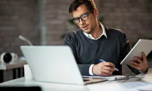 Mężczyzna w okularach pracuje przy laptopie i tablecie, notując. To obraz jego codzienności w **samozatrudnieniu**.
