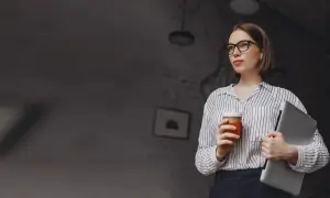 Kobieta w okularach trzymająca kawę i laptopa.