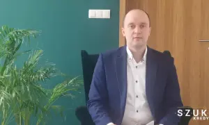 Mężczyzna w garniturze siedzi w fotelu obok rośliny, na tle zielonej ściany i drewnianych drzwi.