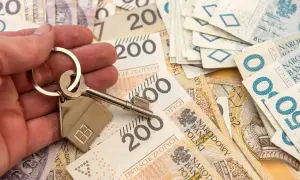 Dłoń trzyma klucz z breloczkiem w kształcie domu na tle polskich banknotów. Obniżka stopy procentowej może oznaczać tańsze kredyty hipoteczne.