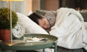 Chory mężczyzna śpi pod kołdrą. Obok łóżka stoi stolik z lekami, termometrem i szklanką wody. To obrazek, który przypomina o ważności ubezpieczenia chorobowego.