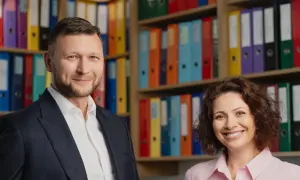 Uśmiechnięci mężczyzna w garniturze i kobieta w różowej koszuli na tle regałów z kolorowymi segregatorami.