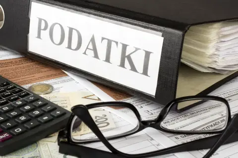 Segregator z napisem "PODATKI", kalkulator, okulary i dokumenty. Obok leży moneta, symbolizująca potencjalną kwotę zmniejszającą podatek.