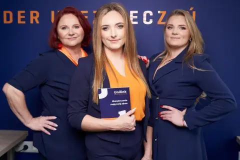 Trzy kobiety w granatowych marynarkach i pomarańczowych bluzkach pozują do zdjęcia. Jedna z nich trzyma broszurę Unilink.