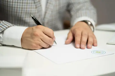 Mężczyzna w garniturze pisze długopisem na białej kartce papieru.