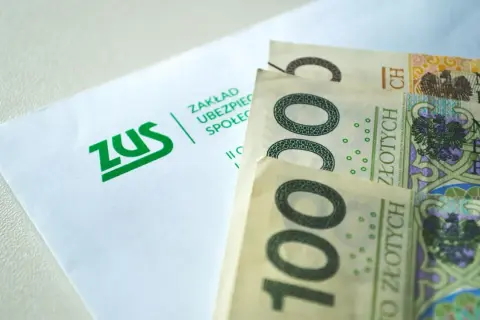 ZUS i polskie banknoty 100 zł. Pieniądze leżą obok koperty z logo ZUS, symbolizując finanse i ubezpieczenia społeczne.