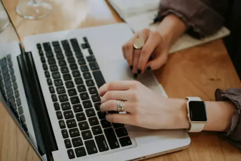 Kobieta pisze na laptopie, analizując odsetki ustawowe. Na palcach ma obrączkę i pierścionek, a na nadgarstku smartwatch.