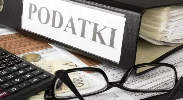 Segregator z napisem "PODATKI", kalkulator, okulary i dokumenty. Obok leży moneta, symbolizująca potencjalną kwotę zmniejszającą podatek.