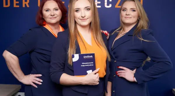 Trzy kobiety w granatowych marynarkach i pomarańczowych bluzkach pozują do zdjęcia. Jedna z nich trzyma broszurę Unilink.