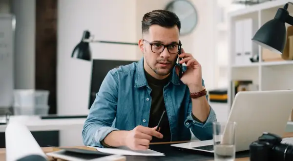Mężczyzna w okularach rozmawia przez telefon przy biurku z laptopem i aparatem.