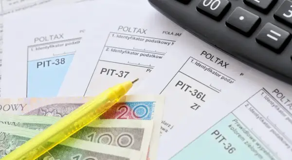 Formularze PIT, polskie banknoty, żółty długopis i kalkulator.