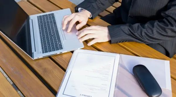 Biznesmen pracuje na laptopie przy drewnianym stole, obok dokumenty z nagłówkiem "CONTRACT". Przygotowuje wpis do księgi wieczystej.