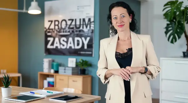 Kobieta w beżowej marynarce, obok niej na biurku leży tablet i notatnik, który może służyć jako księga przychodów i rozchodów.