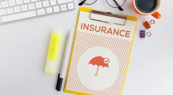 Planowanie ubezpieczenia zdrowotnego bez pracy. Na biurku klawiatura, okulary, kawa i dokument "INSURANCE" z parasolką.