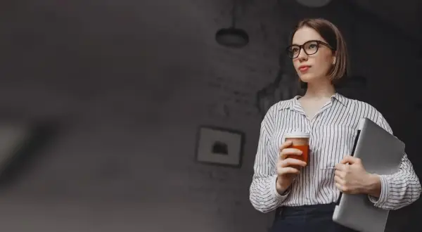 Kobieta w okularach trzymająca kawę i laptopa.