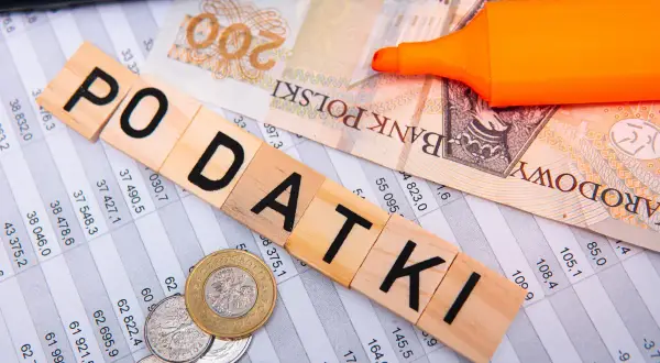 Drewniane klocki z napisem "PODATKI", banknot 200 zł, monety i pomarańczowy marker na arkuszu kalkulacyjnym. Myśląc o przyszłości, warto pamiętać o podatku katastralnym.