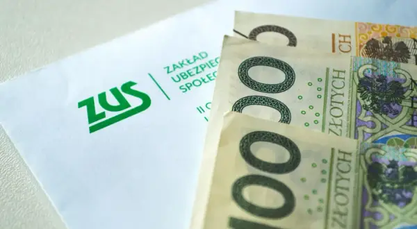 ZUS i polskie banknoty 100 zł. Pieniądze leżą obok koperty z logo ZUS, symbolizując finanse i ubezpieczenia społeczne.