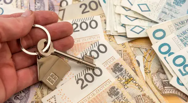 Dłoń trzyma klucz z breloczkiem w kształcie domu na tle polskich banknotów. Obniżka stopy procentowej może oznaczać tańsze kredyty hipoteczne.