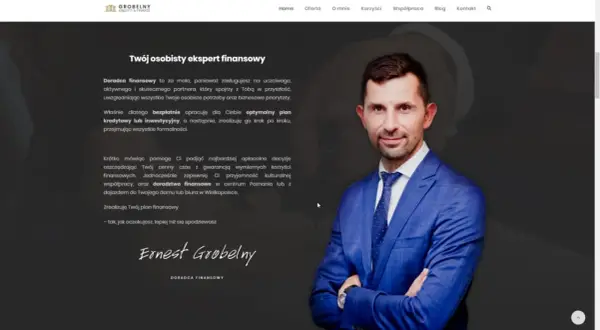 Mężczyzna w niebieskiej marynarce z krawatem, uśmiecha się, stojąc na tle strony internetowej.