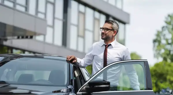 Biznesmen w białej koszuli i krawacie opiera się o drzwi czarnego samochodu. Czeka na niego kolejna kilometrówka.