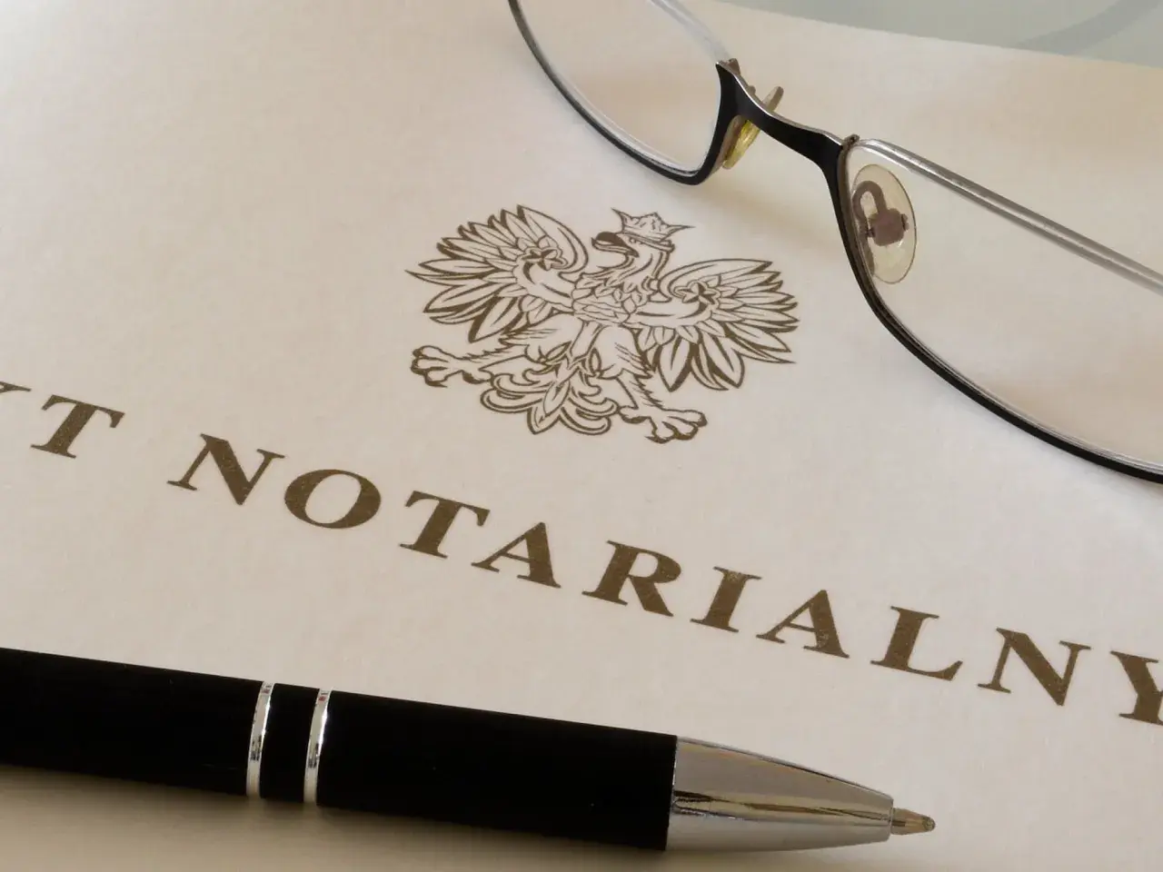 Okulary i długopis leżą na dokumencie z godłem Polski i napisem "AKT NOTARIALNY". To dokument, który wymaga opłaty, czyli taksa notarialna.