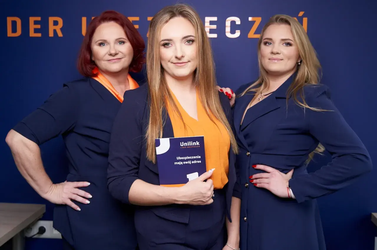 Trzy kobiety w granatowych marynarkach i pomarańczowych bluzkach pozują do zdjęcia. Jedna z nich trzyma broszurę Unilink.