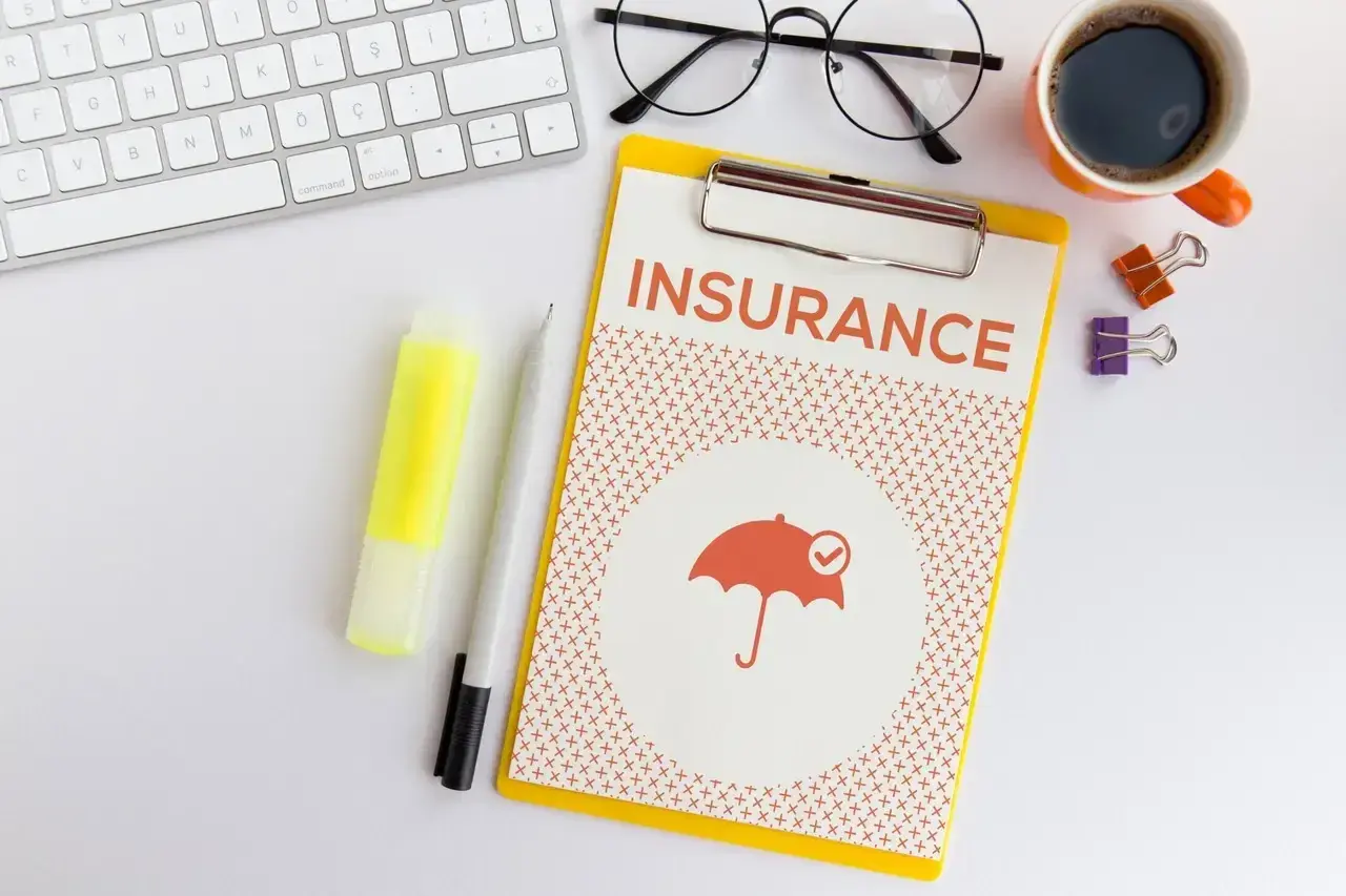 Planowanie ubezpieczenia zdrowotnego bez pracy. Na biurku klawiatura, okulary, kawa i dokument "INSURANCE" z parasolką.