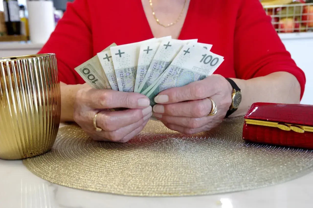 Dłonie trzymające banknoty 100 zł. Zastanawiasz się, komu przysługuje przeliczenie emerytury? Obok złoty wazon i czerwona szkatułka.