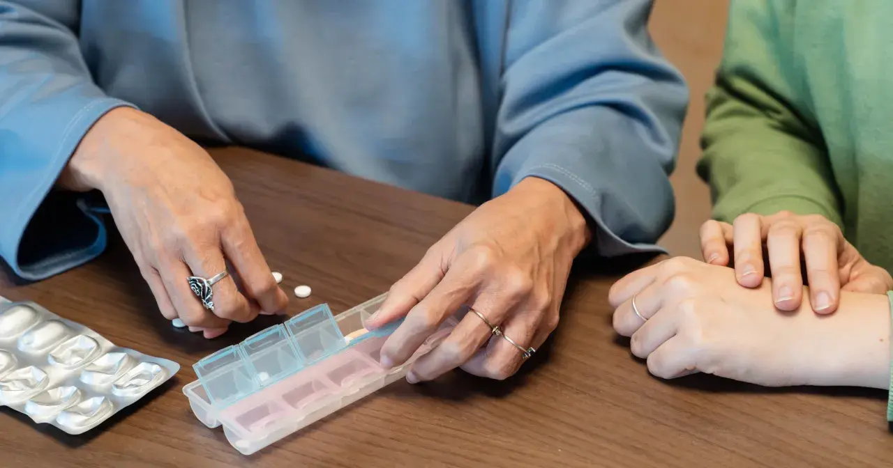 Dwie osoby sortują tabletki w pojemniku na leki. Dostępne są darmowe leki dla seniorów.