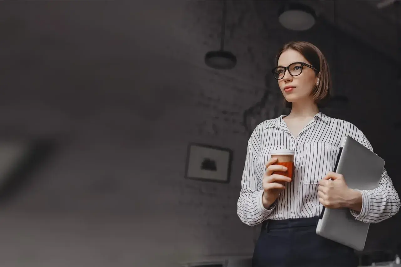 Kobieta w okularach trzymająca kawę i laptopa.