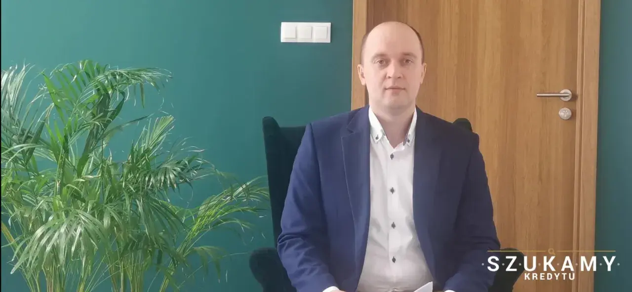 Mężczyzna w garniturze siedzi w fotelu obok rośliny, na tle zielonej ściany i drewnianych drzwi.