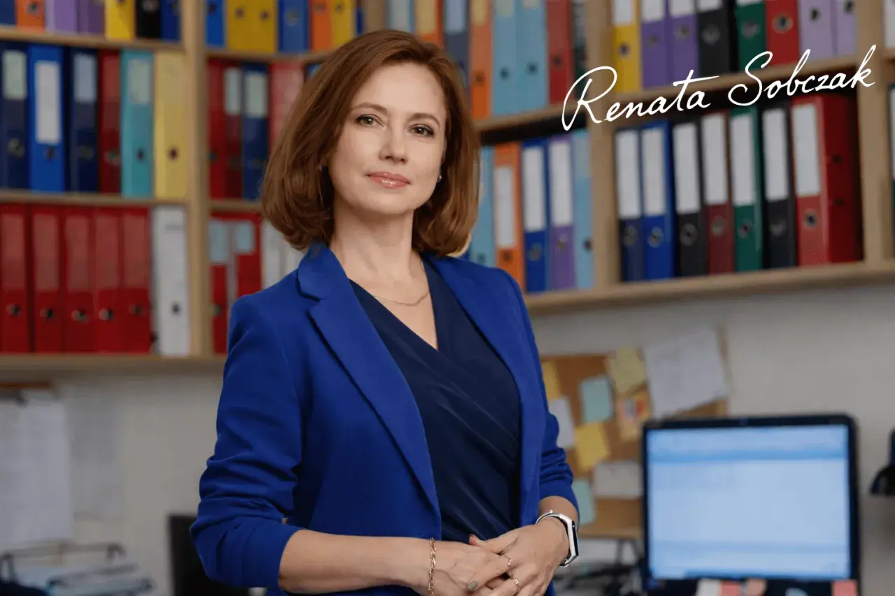 Kobieta w niebieskiej marynarce stoi przed regałami z kolorowymi segregatorami.