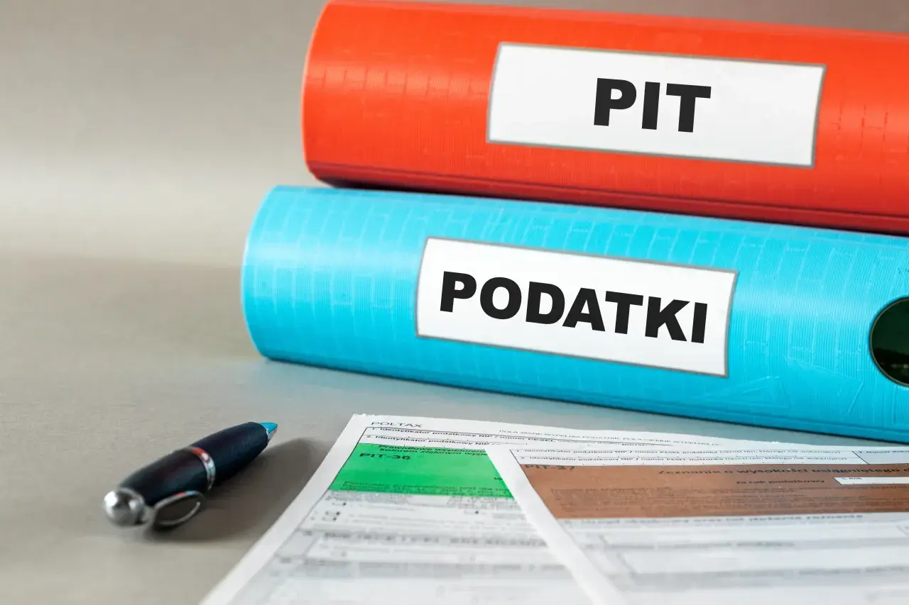 Dwa segregatory, jeden czerwony z napisem "PIT", drugi niebieski z napisem "PODATKI", leżą na dokumentach podatkowych.