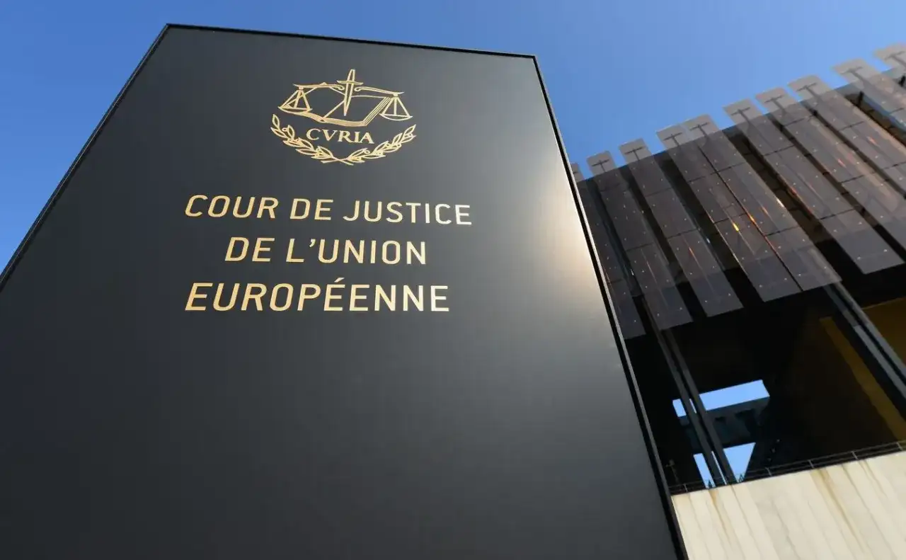 Czarny szyld z napisem "COUR DE JUSTICE DE L'UNION EUROPÉENNE" i symbolem wagi. W tle nowoczesny budynek.