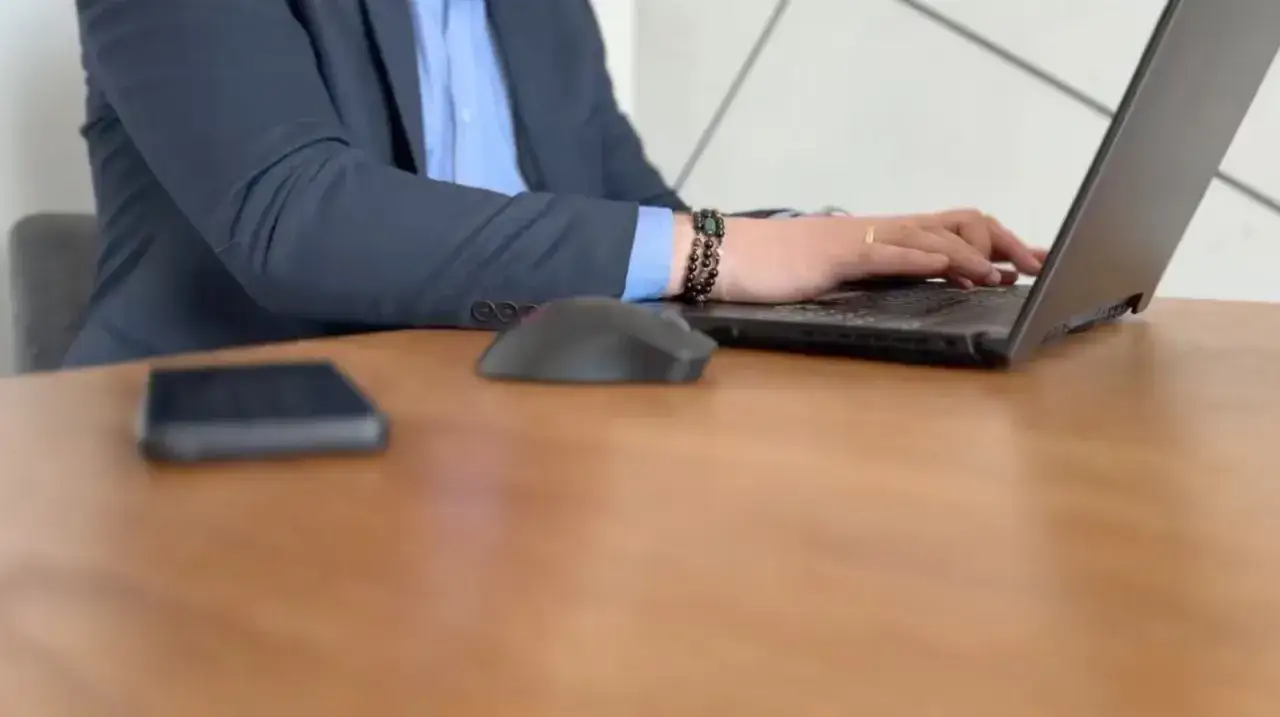 Osoba w garniturze pracuje na laptopie, obok leży myszka i telefon.
