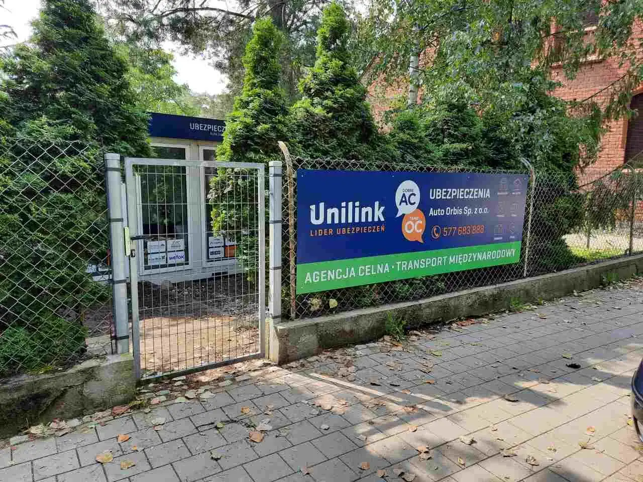 Brama z siatki przed budynkiem z banerem Unilink i informacją o ubezpieczeniach.