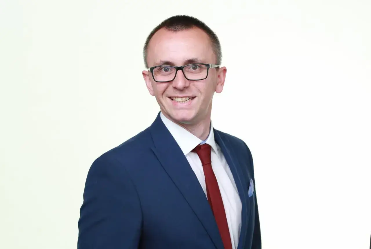 Mężczyzna w okularach, granatowej marynarce i czerwonym krawacie, uśmiecha się na białym tle.