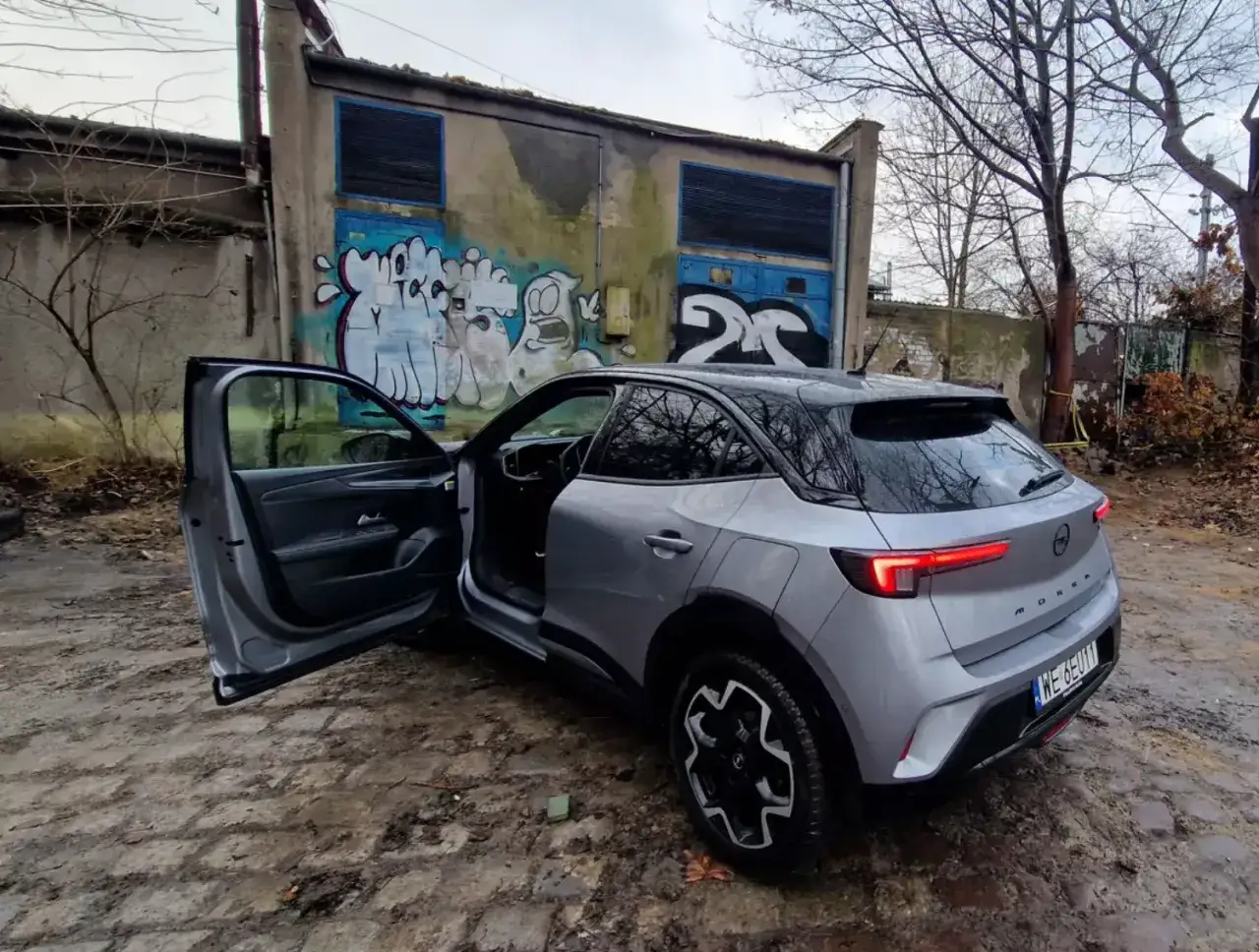 Srebrny Opel Mokka z otwartymi drzwiami, gotowy do darowizny samochodu po leasingu. Tło to zniszczony budynek z graffiti.