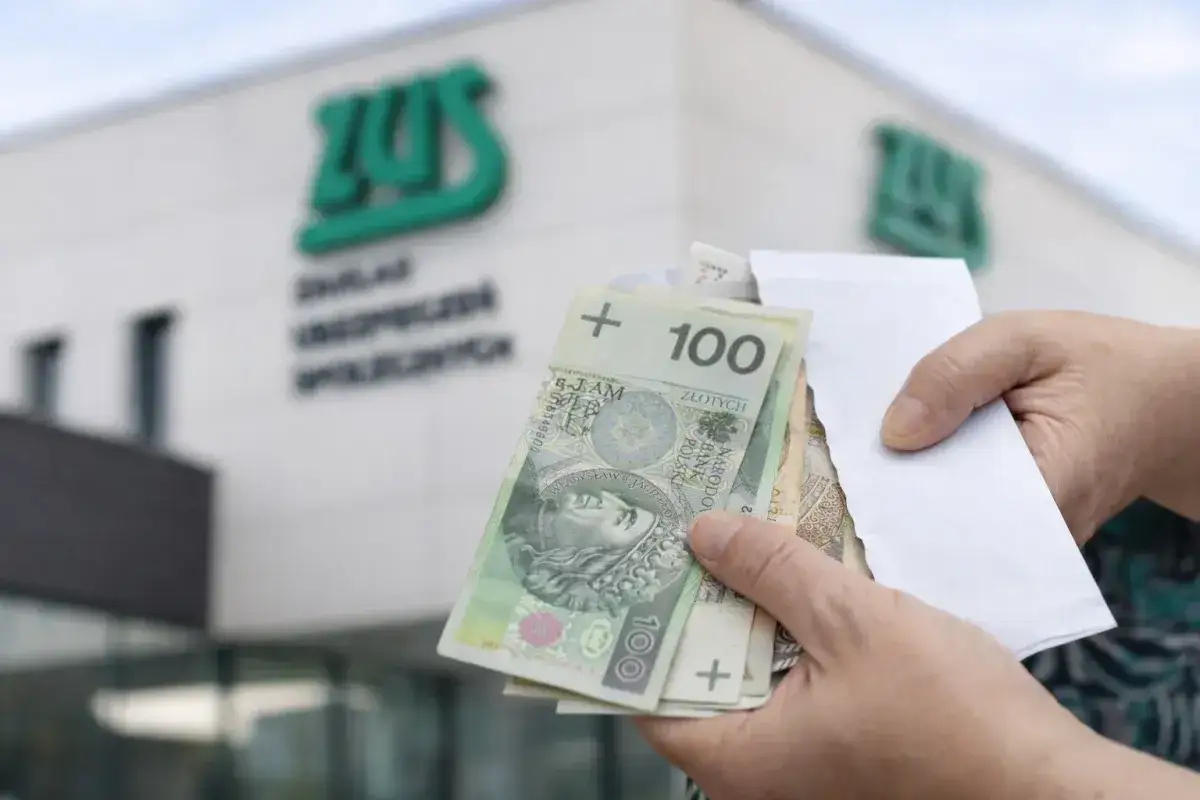 Ręce trzymają polskie banknoty przed budynkiem ZUS. Sprawdzanie 14 emerytury tabela netto.