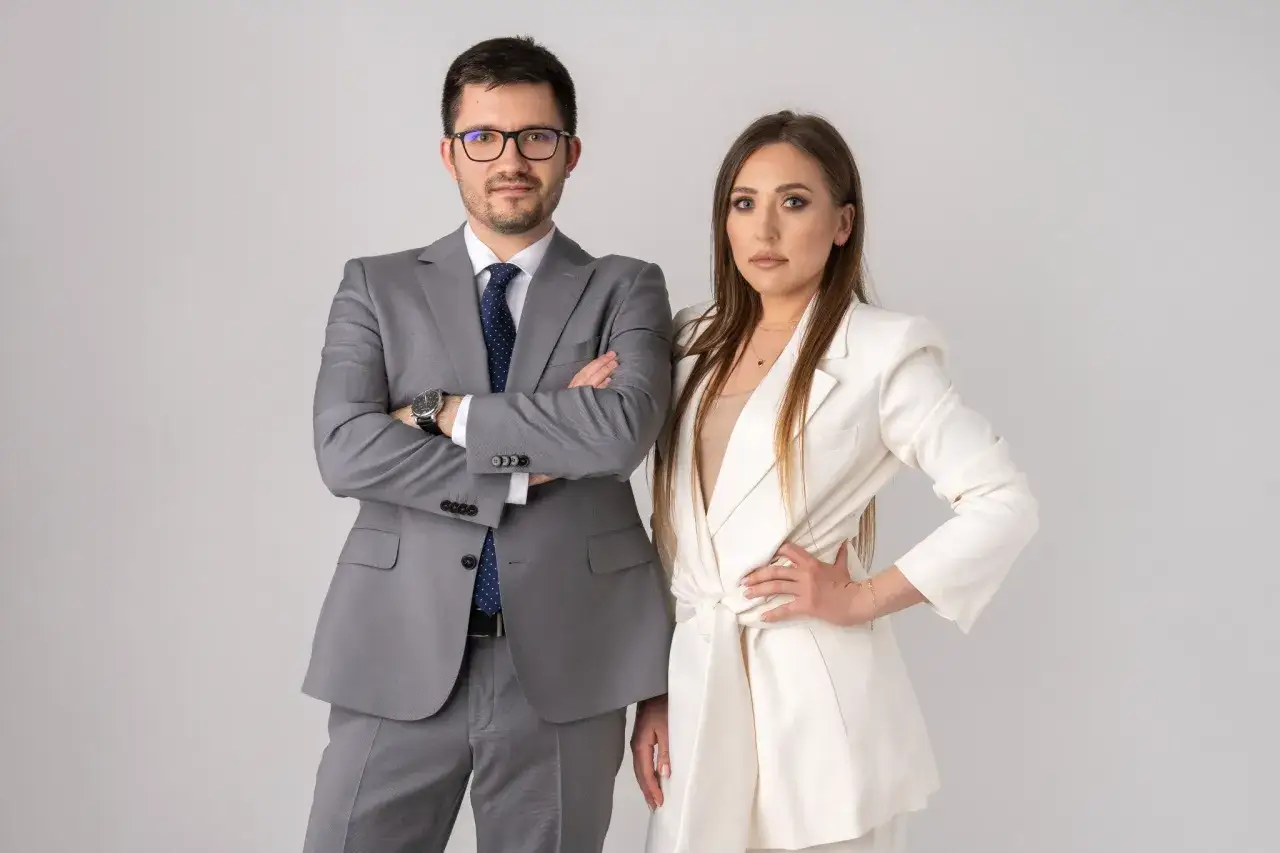 Para w garniturach, mężczyzna z założonymi rękami, kobieta z ręką na biodrze.