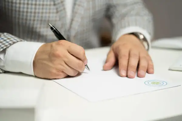 Mężczyzna w garniturze pisze długopisem na białej kartce papieru.