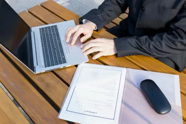 Biznesmen pracuje na laptopie przy drewnianym stole, obok dokumenty z nagłówkiem "CONTRACT". Przygotowuje wpis do księgi wieczystej.